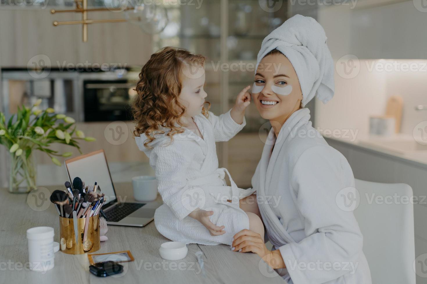 glad ung mamma lärare sin lilla dotter att göra makeup. culry liten flicka applicerar skönhetsplåster under ögonen på mamma använder skönhetsprodukter naturl kosmetika. hudvård och familjetid koncept foto