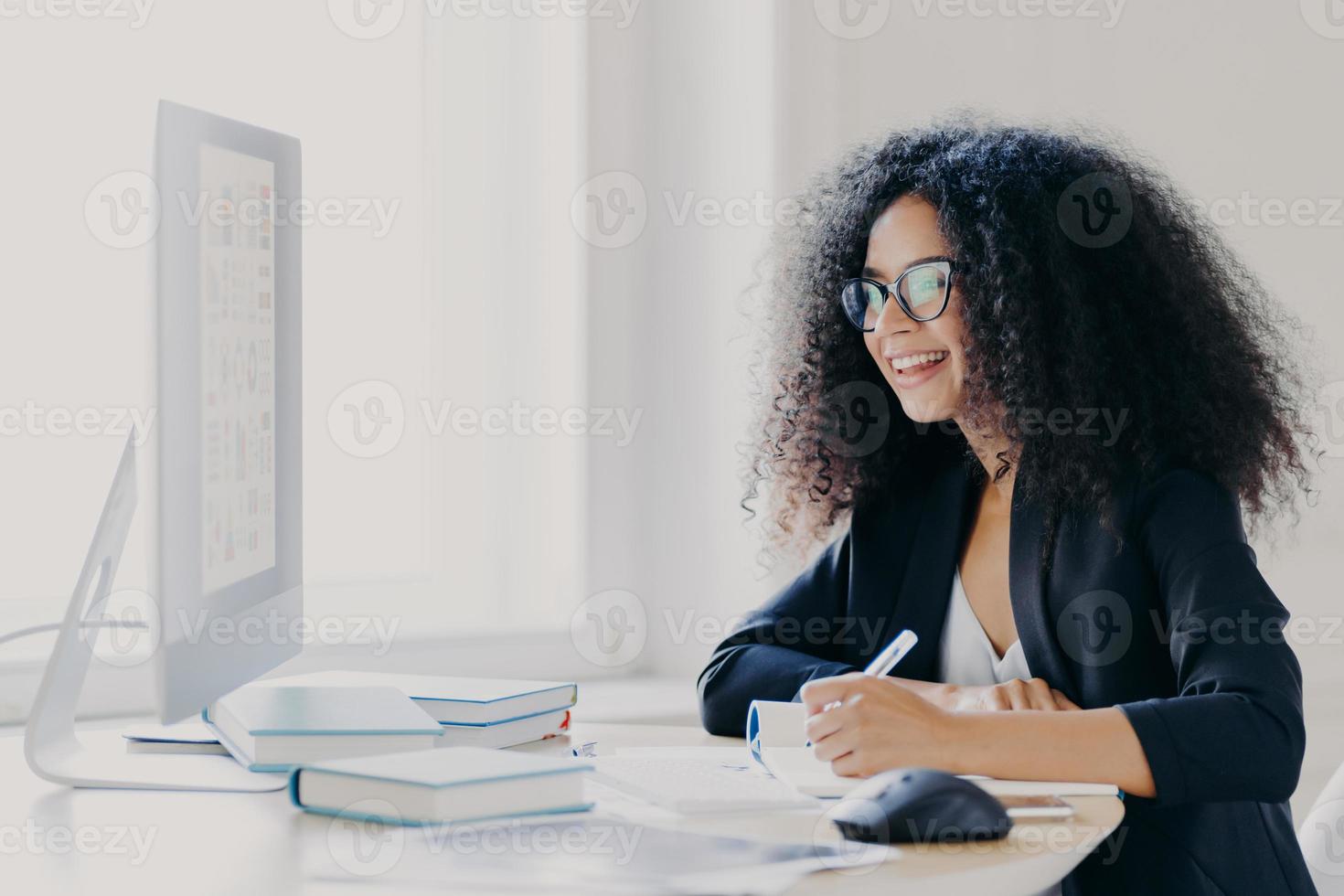 professionell kvinna med lockigt hår gör en rapport, fokuserar på skärmen, skriver ner information, bär glasögon och högtidsdräkt, noterar idén för startup-planering, poserar i kontorets interiör. foto