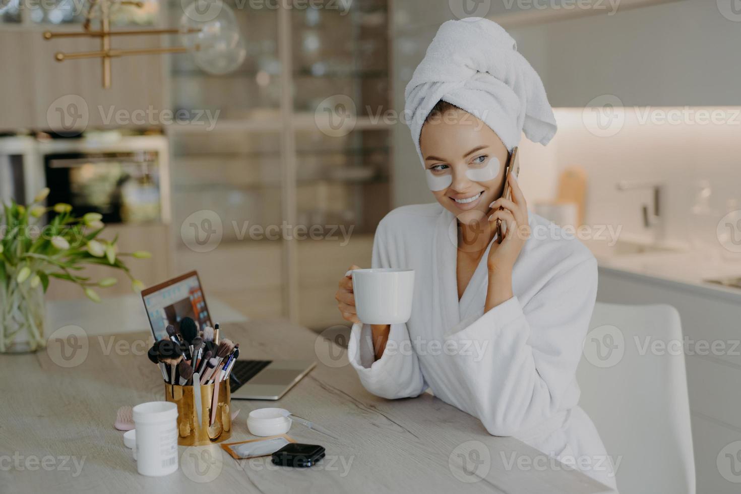 glad ung europeisk kvinna genomgår skönhetsbehandlingar efter morgondusch bär hydrogel fuktgivande plåster klädd i vit morgonrock dricker aromatiskt kaffe samtal via mobiltelefon poserar inomhus foto