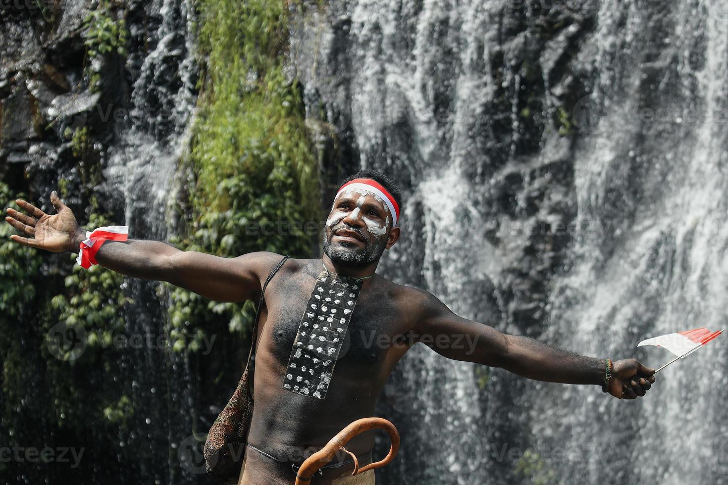 uttrycksfull papua man bär traditionella kläder av dani stam, röd-vit pannband och armring håller lilla Indonesiska flaggan och firar Indonesiens självständighetsdag mot vattenfall bakgrund. foto