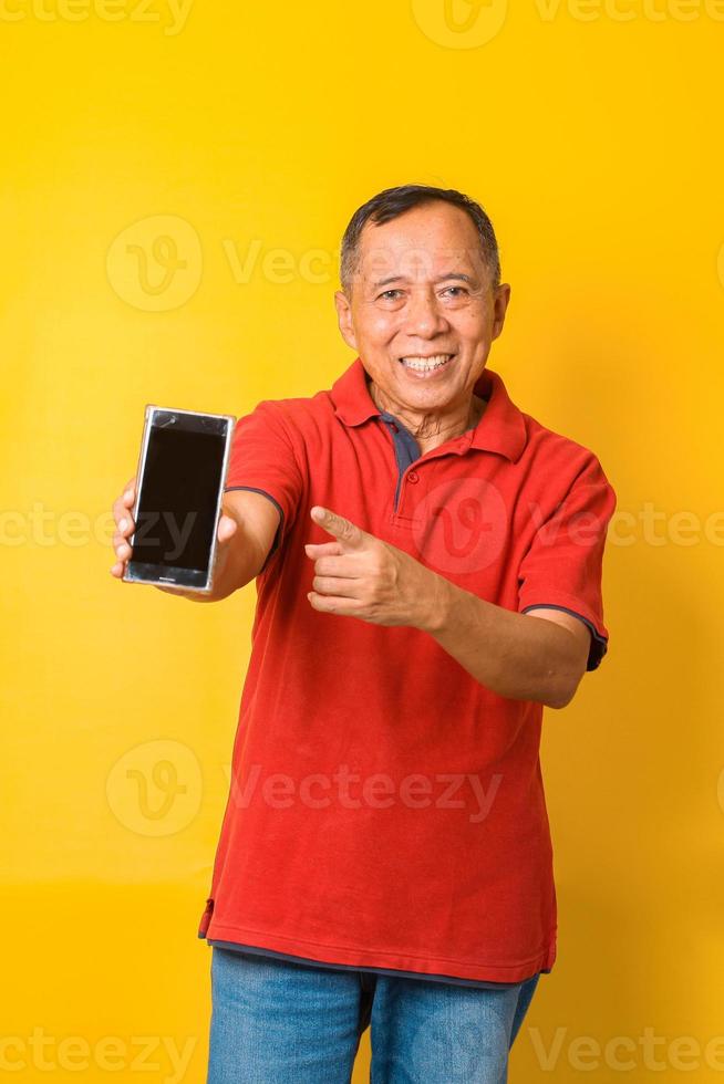 foto av asiatisk gammal man som håller en tom telefon med tom skärm med direkt finger som pekar på skärmen iförd röd t-shirt isolerad på gul bakgrund