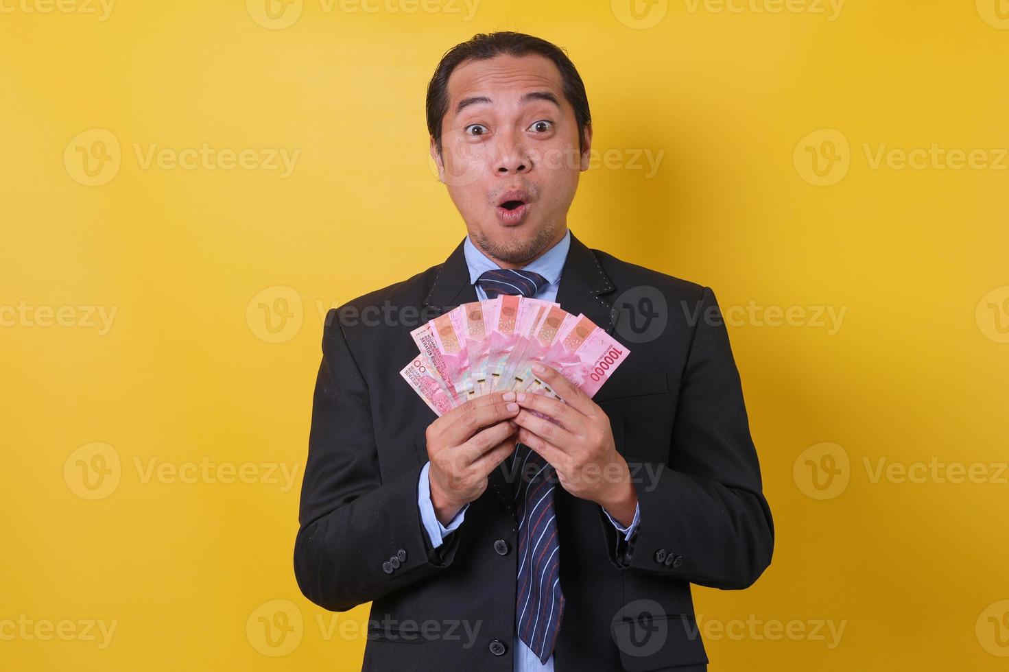 asiatisk affärsman i kostym står mot gul bakgrund, visar pengar hundra tusen rupiah på händer med wow uttryck. finansiellt och sparande koncept. foto