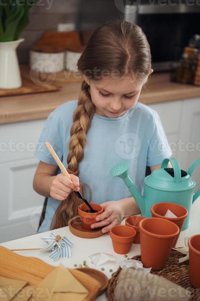 liten flicka som sitter vid bordet hemma och sår frön i blomkrukor. foto