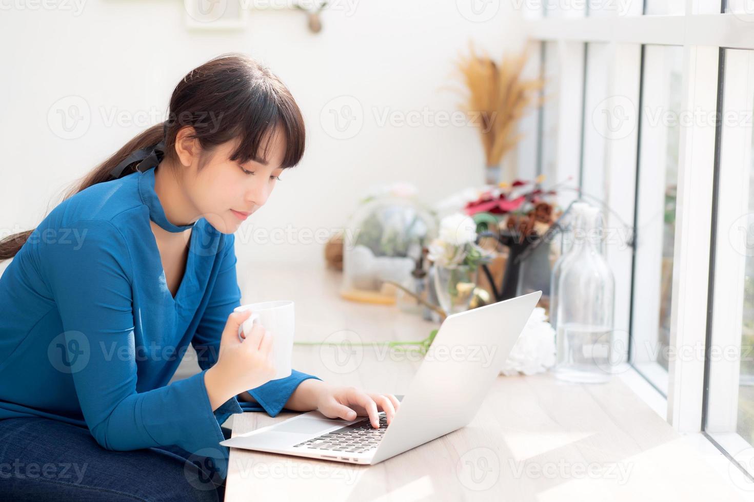 vacker ung frilansande asiatisk kvinna som ler arbetar och på bärbar dator vid skrivbordet kafé med professionell, asiatisk tjej som använder anteckningsbok och dricker kaffe, affärs- och livsstilskoncept. foto