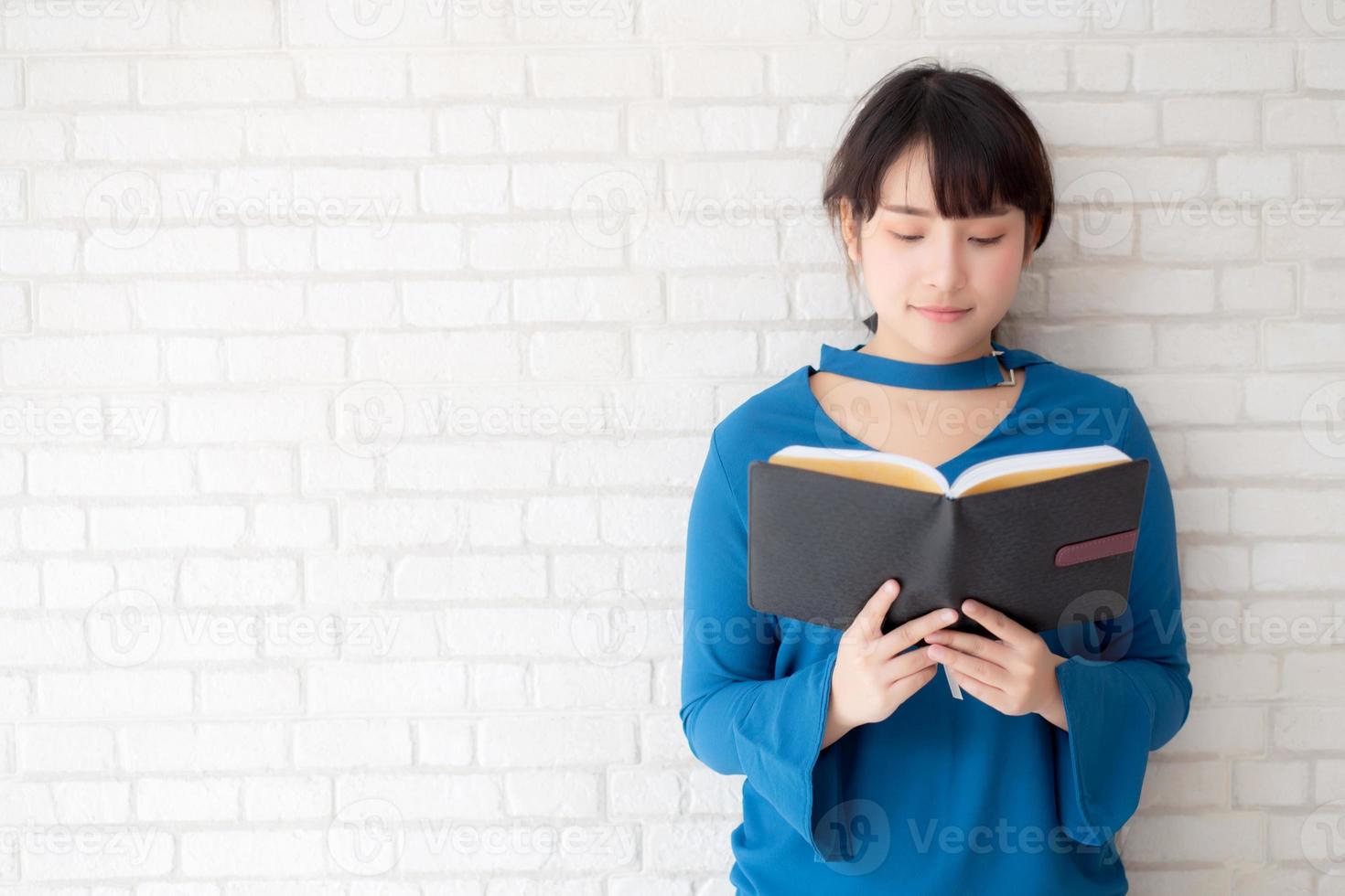 vacker av porträtt ung asiatisk kvinna lycka koppla av stående läsbok på betong cement vit bakgrund, flicka glad studieinnehåll litteratur, utbildning och livsstilskoncept. foto