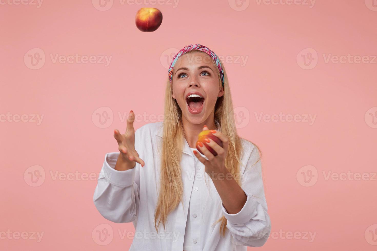 glad söt dam med långt blont hår som jonglerar med persikor i studion, har kul över rosa bakgrund, bär färgat pannband och vit skjorta foto