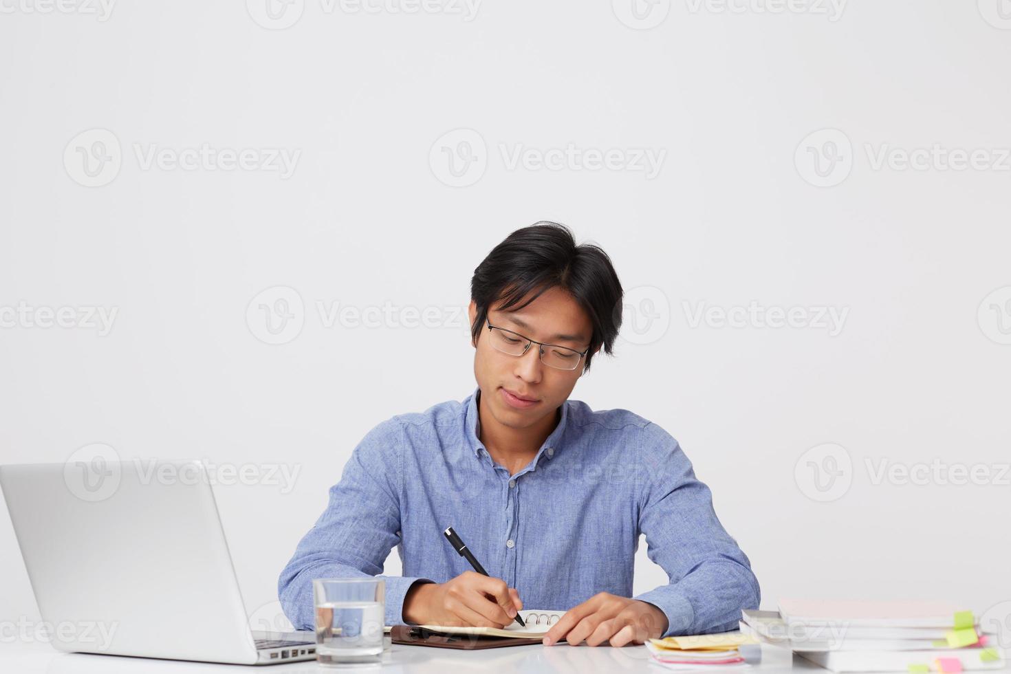 tankeväckande fokuserad asiatisk ung affärsman i glasögon som arbetar vid bordet med laptop och skrivplan i anteckningsbok över vit bakgrund foto