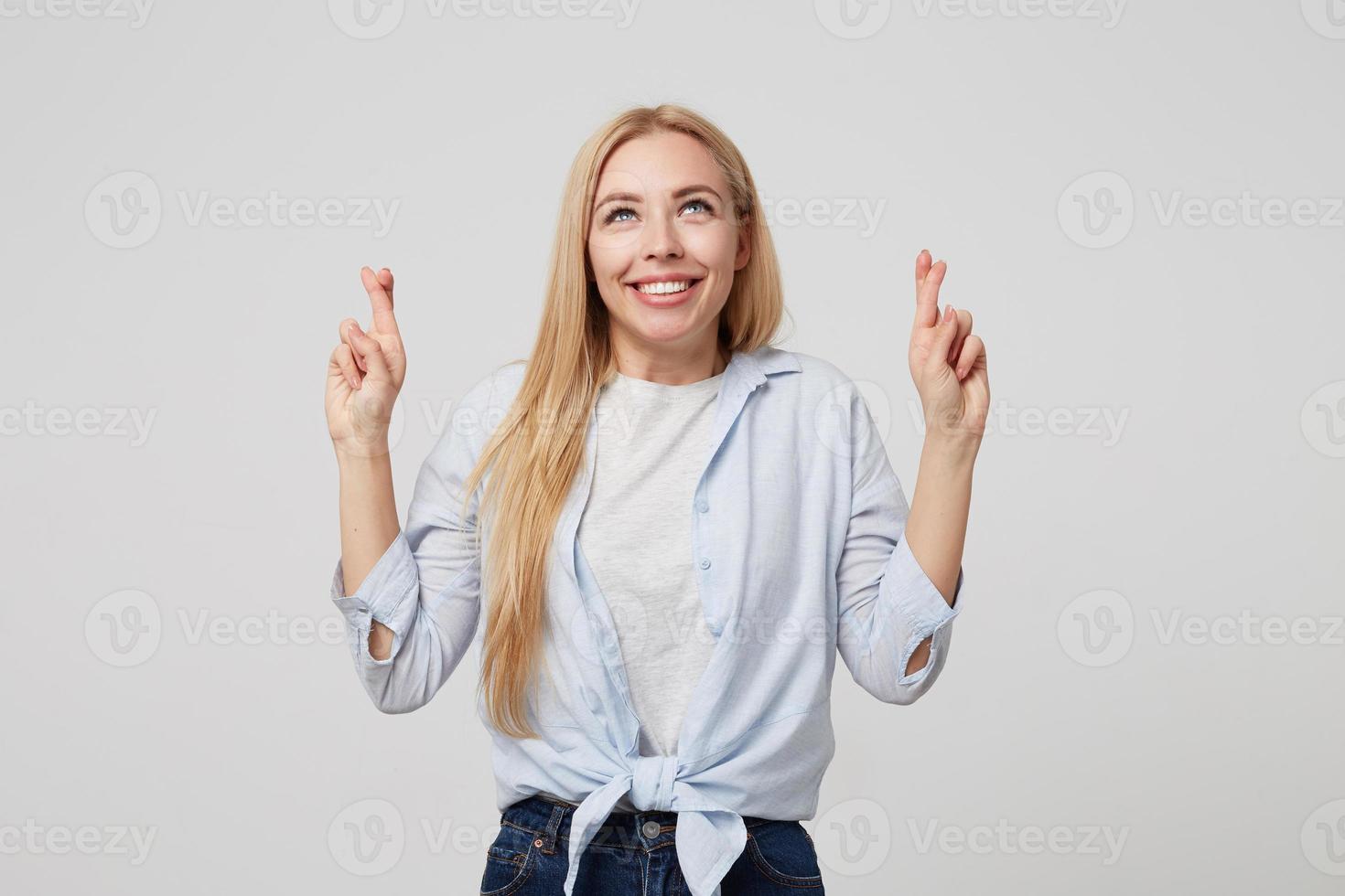 inomhusbild av en leende ung flicka med långt blont hår klädd i blå skjorta och jeans, som står över vit bakgrund, håller tummarna för lycka och tittar uppåt foto