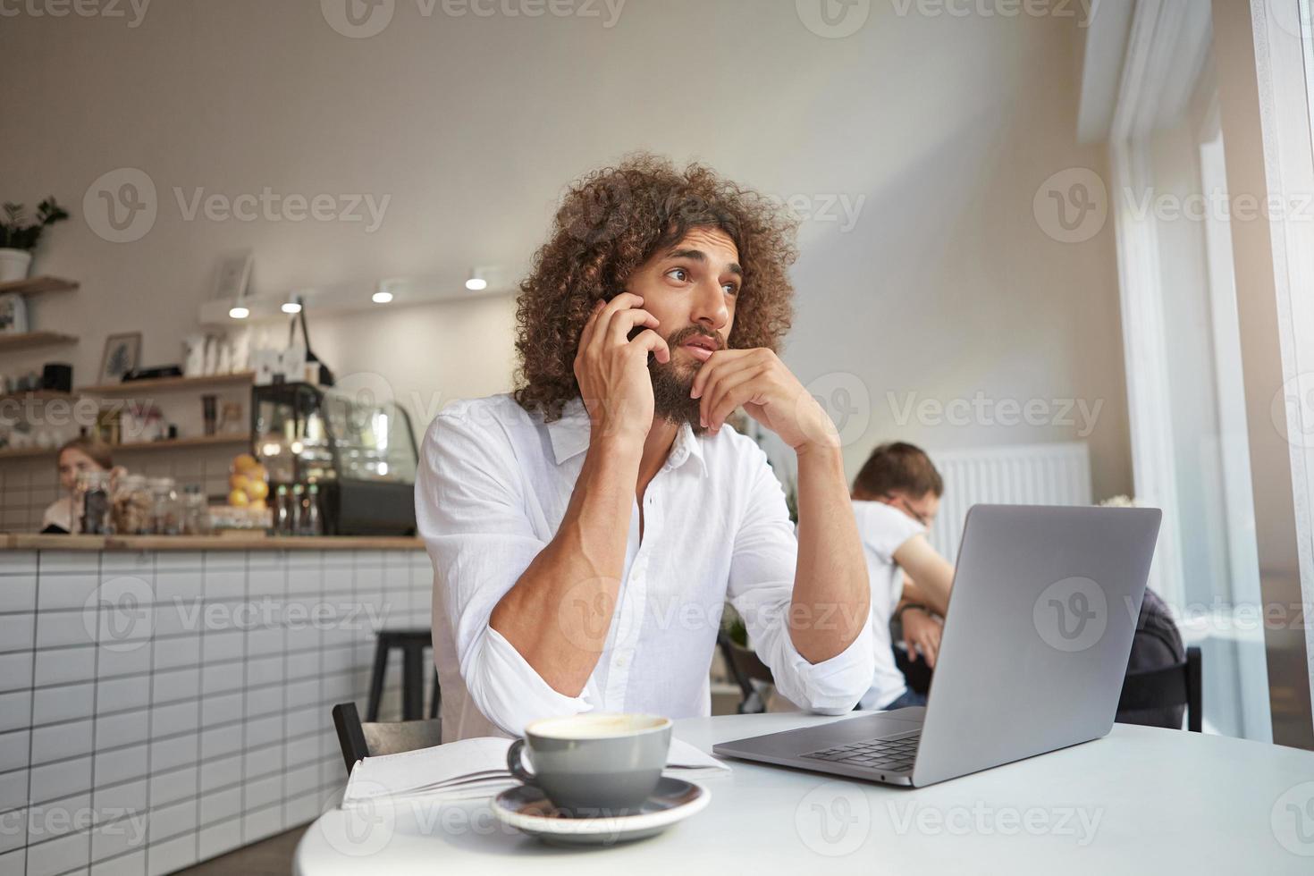 inomhusbild av stilig söt ung man som arbetar på distans på offentlig plats, pratar i telefon och tittar eftertänksamt i fönstret, håller hakan med handen och rynkig panna foto