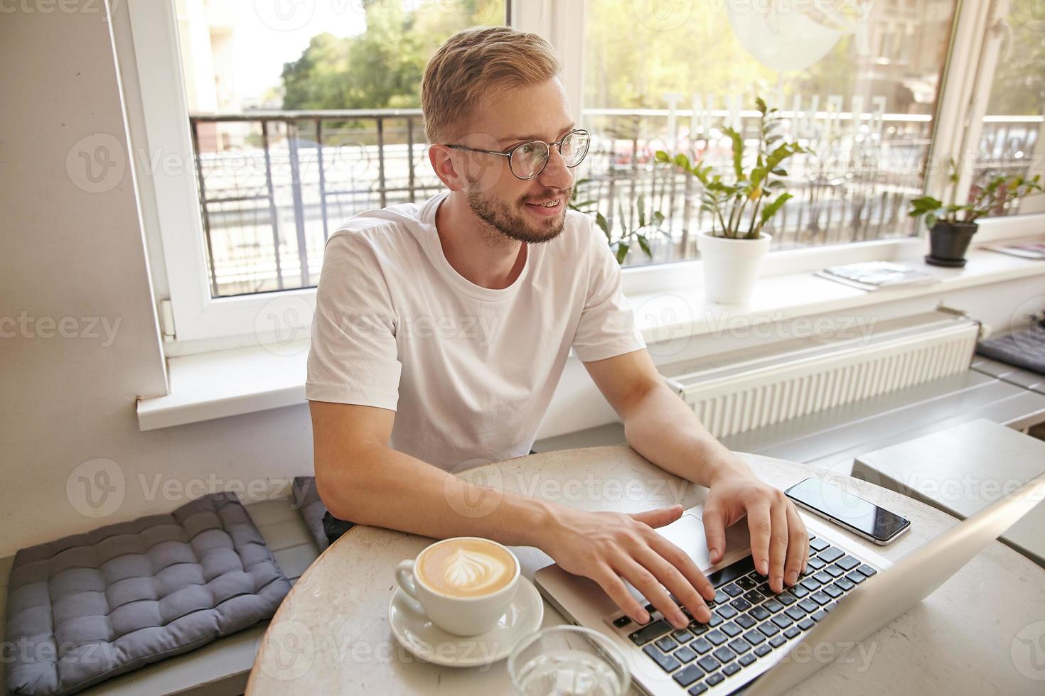 porträtt av ung söt kille, som arbetar på distans på stadskaféet, tittar bort med ett mjukt leende, gör paus med jobbet och tänker på något positivt foto