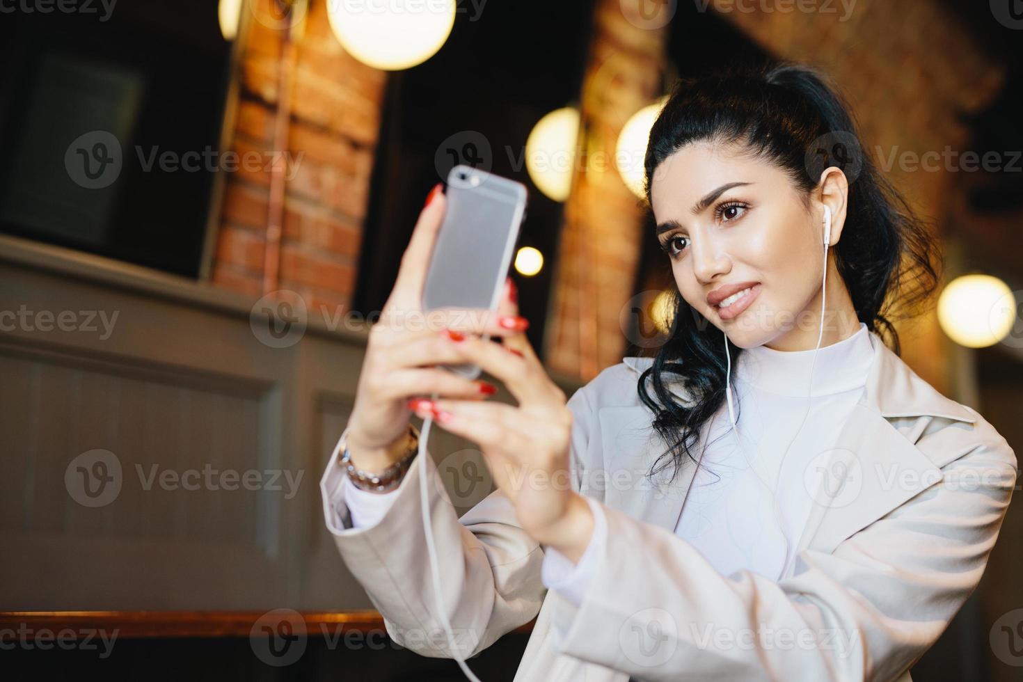 vacker kvinna med mörkt hår bundet i hästsvans gör selfie medan du sitter på café med sin smartphone. välvårdad kvinna i eleganta kläder tittar på skärmen på hennes telefon som gör foto