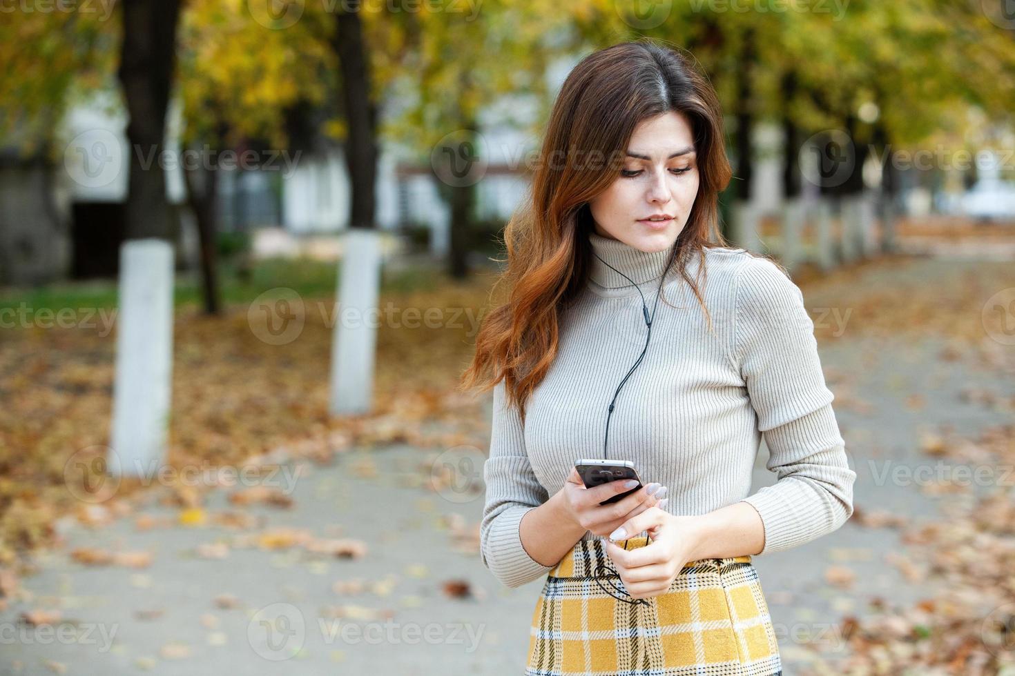 bild av vacker elegant kvinna med mobiltelefon. ung europeisk flicka står på gatan och använder mobiltelefon. kvinna som lyssnar på musik med telefonen och har kul. mobilt internet koncept. foto