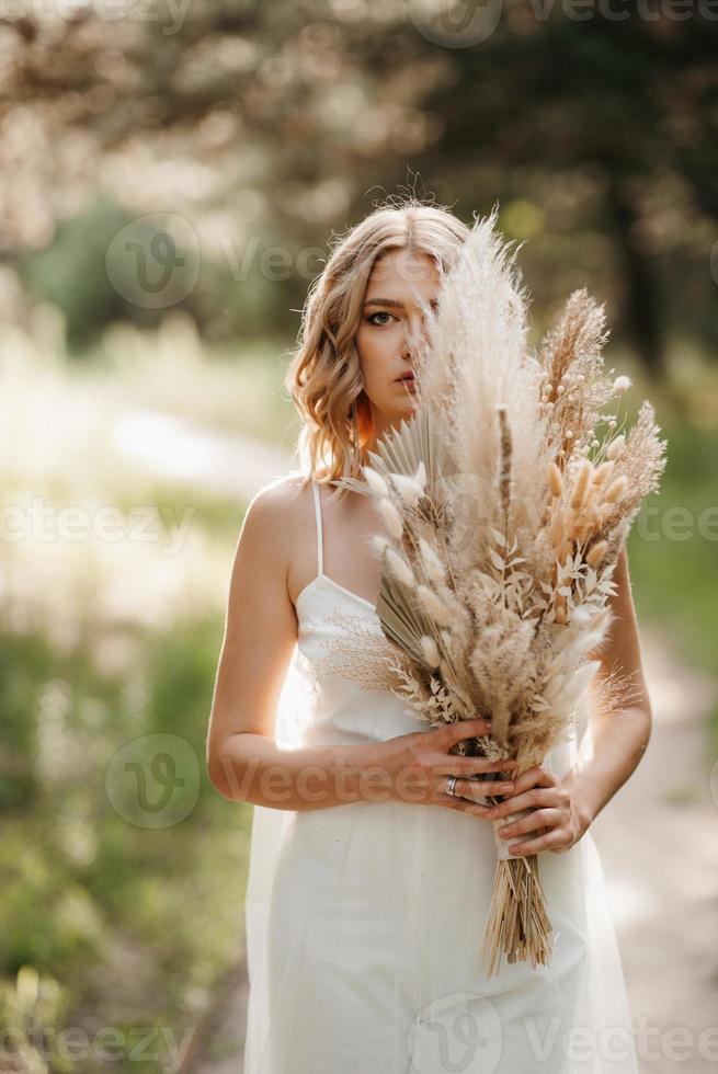 glad brud flicka i en vit ljus klänning med en bukett torkade blommor foto