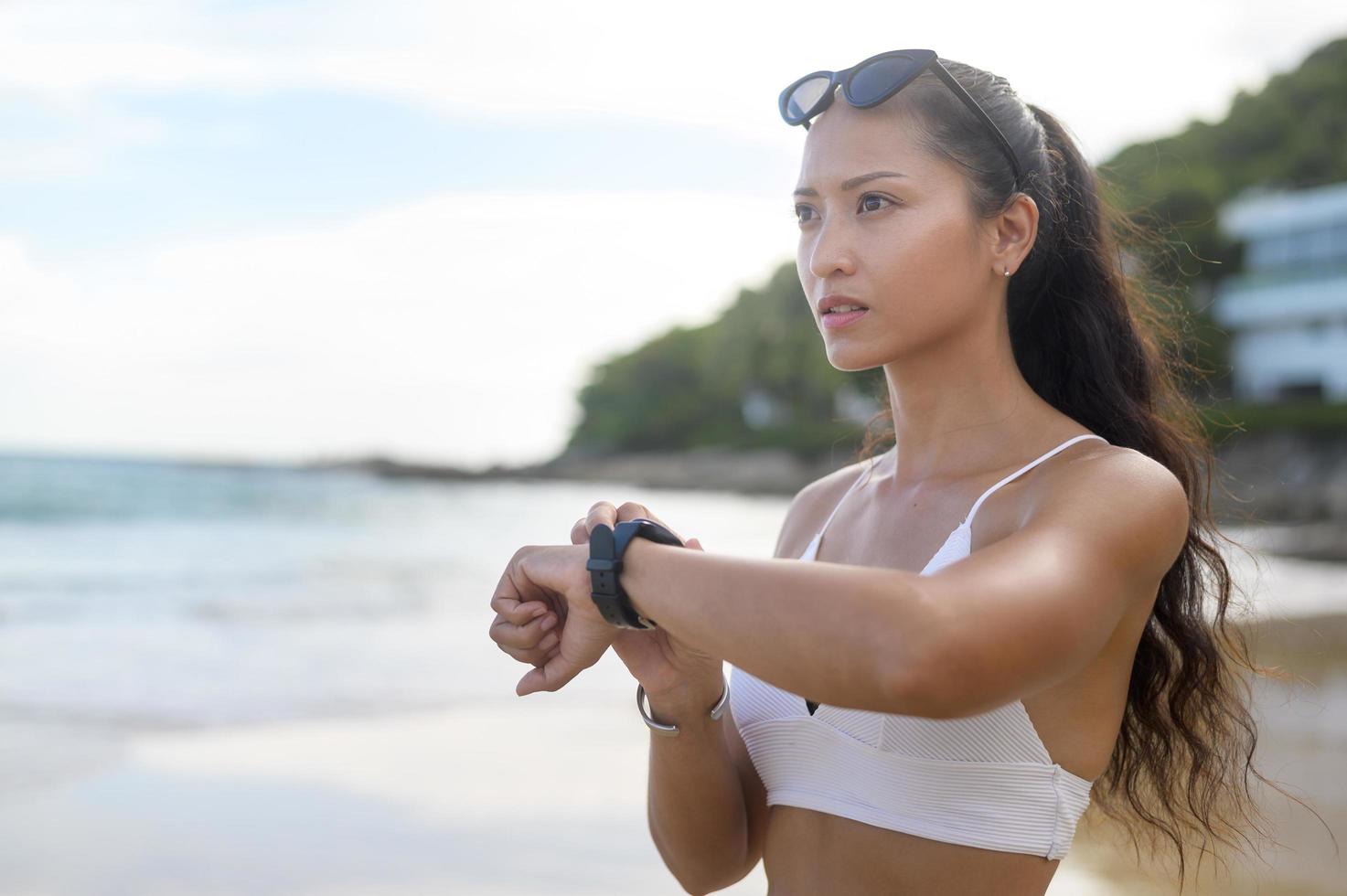 ung vacker kvinna i bikini kopplar av och använder smartwatch på stranden, sommaren, semestern, semestern och hälsokonceptet. foto