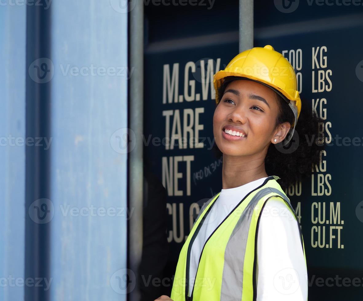 leende vackra afroamerikanska kvinnliga förman arbetare bär gul säkerhetshjälm tittar på lastlådor lastning på containergården, porträtt. industriingenjör kvinna arbetar på internationella varv foto