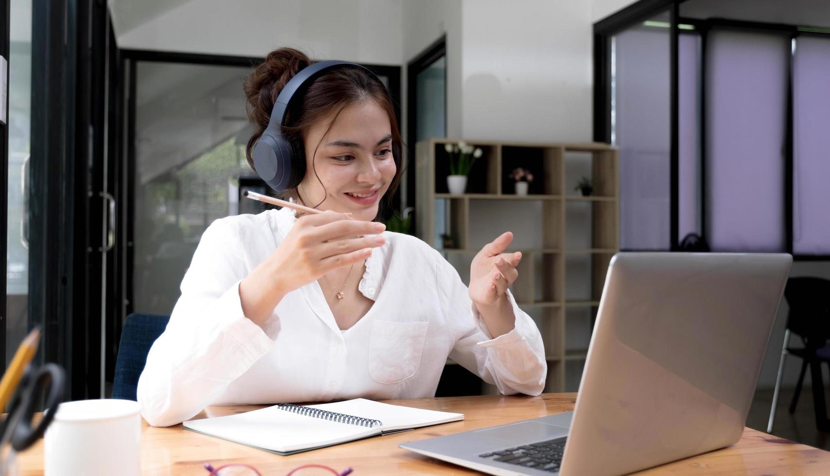 leende ung asiatisk kvinna i headset våg hälsa pratar på webbkamera virtuell konversation på bärbar dator, glad kvinna i trådlösa hörlurar prata i videosamtal på dator, konsultera klient online foto