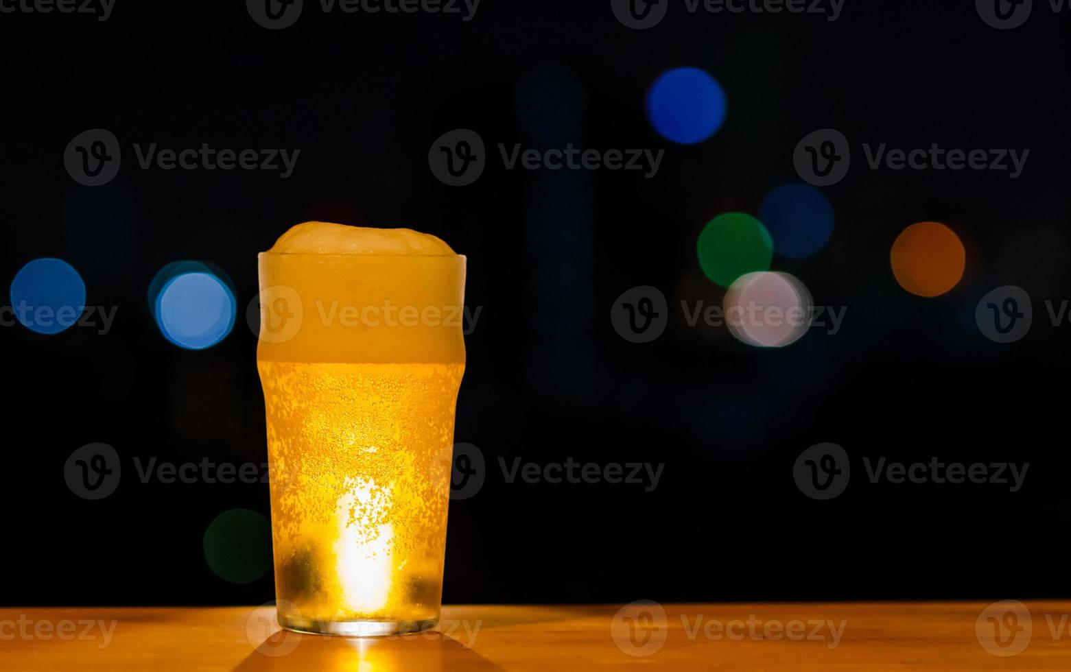 ett glas öl med dess skum sätter på träbord av baren isolerad på mörk natt bakgrund med färgglada bokeh lampor på takbaren. foto