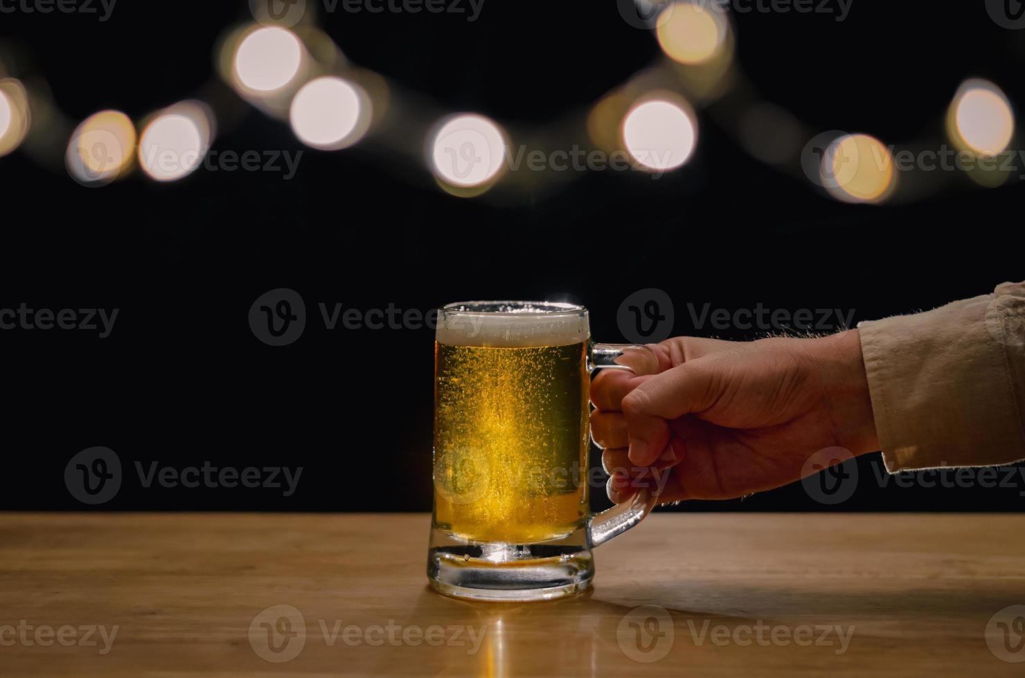 hand som håller ett glas öl på träbord som har bokeh-ljus på toppen med mörk bakgrund. foto