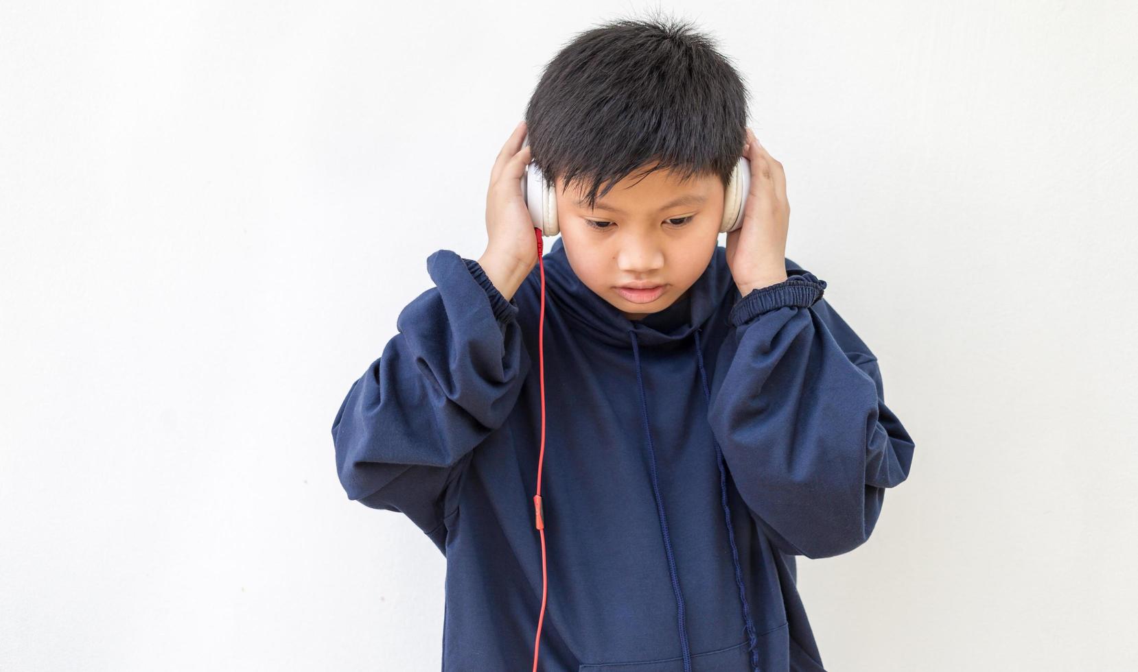 söt asiatisk pojke i en hoodie poserar stående leende avslappnad och glad lyssnar på musik med hörlurar. känslomässigt porträtt av en ung pojke som njuter av musik foto