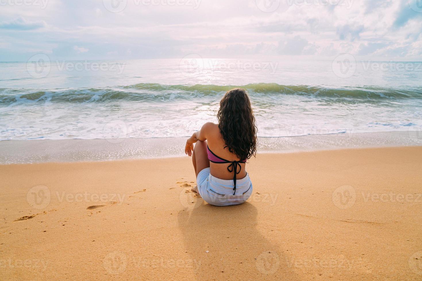 siluett av ung kvinna på stranden. latinamerikansk kvinna sitter på stranden sanden tittar på himlen en vacker sommardag foto