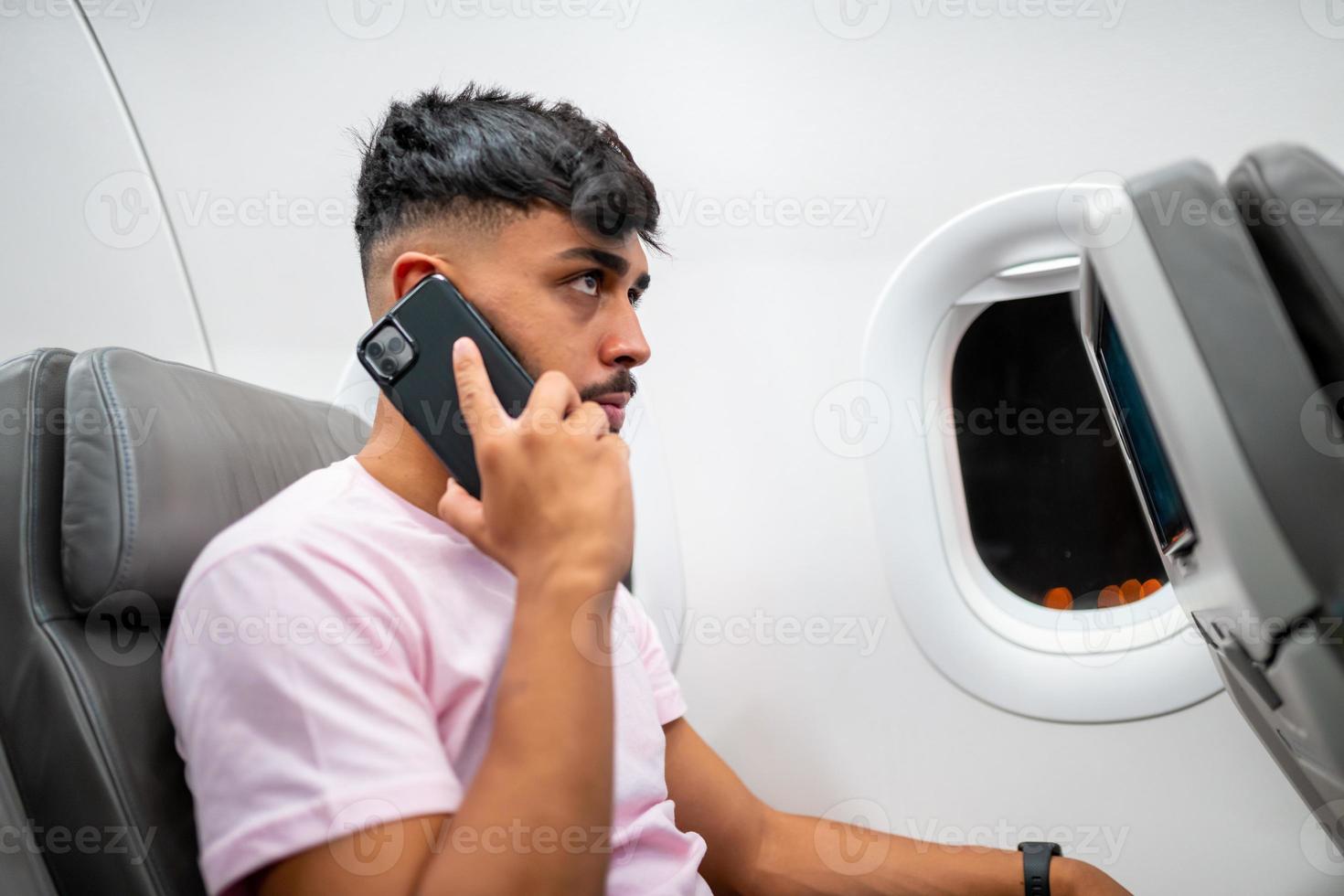Latinamerikansk ung man pratar i mobiltelefon när han sitter på planet nära fönstret. foto