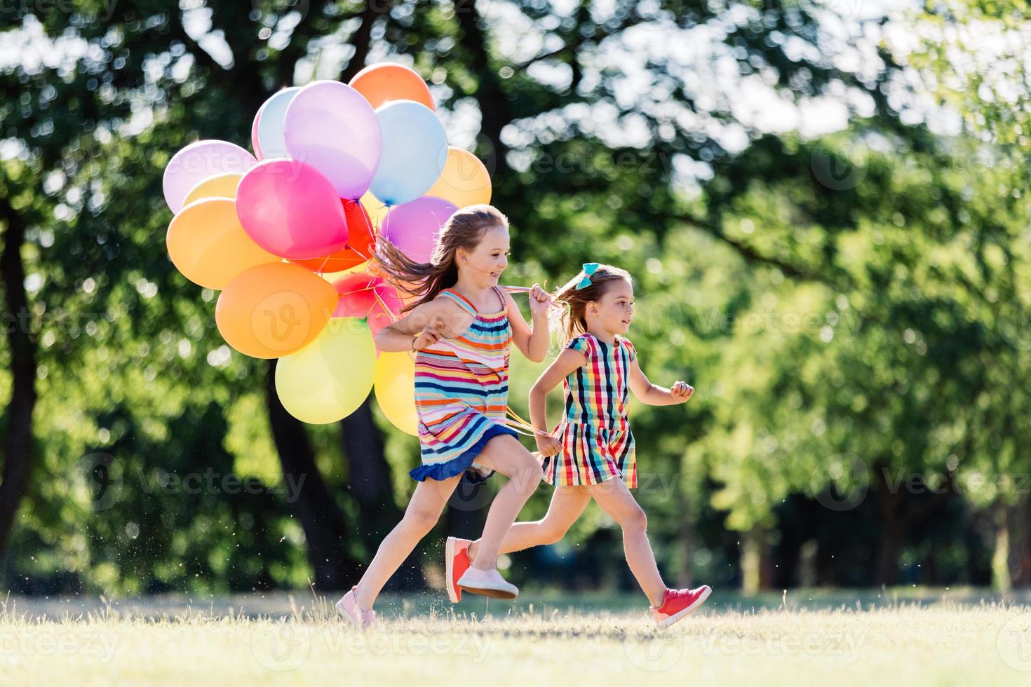 två små glada tjejer springer med ett gäng färgglada ballonger. foto