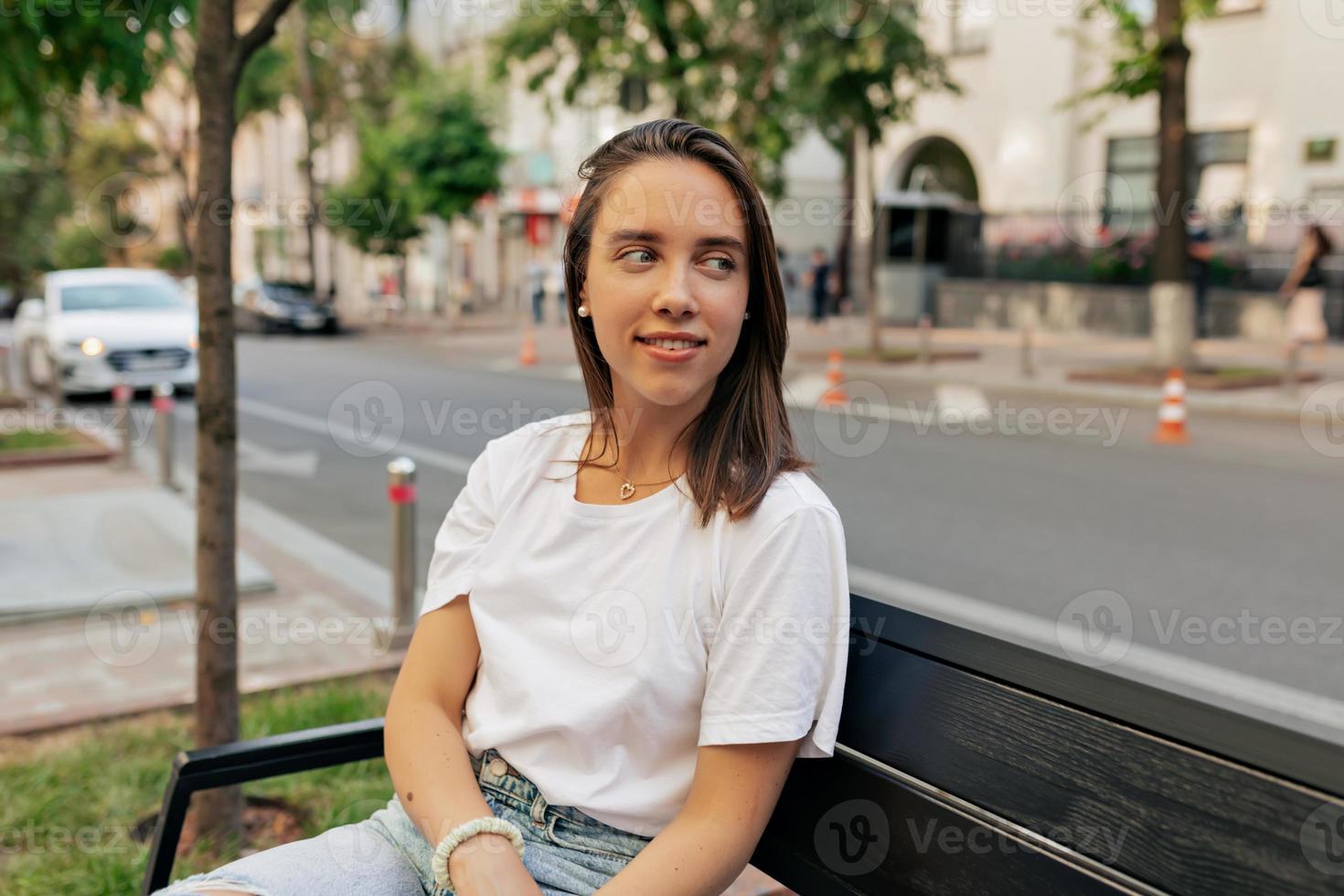 bedårande söt kvinna med mörkt hår i vit t-shirt sitter på bänken på sommargatan i staden i varm vårdag foto