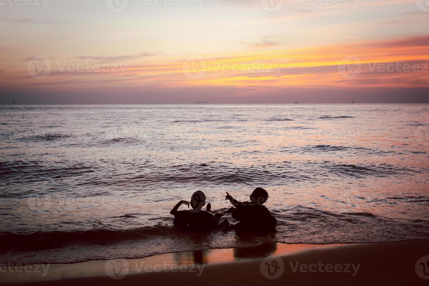 en siluett av två pojkar som slappar i havet vid stranden på kvällen efter solnedgången under skymningshimlen. foto