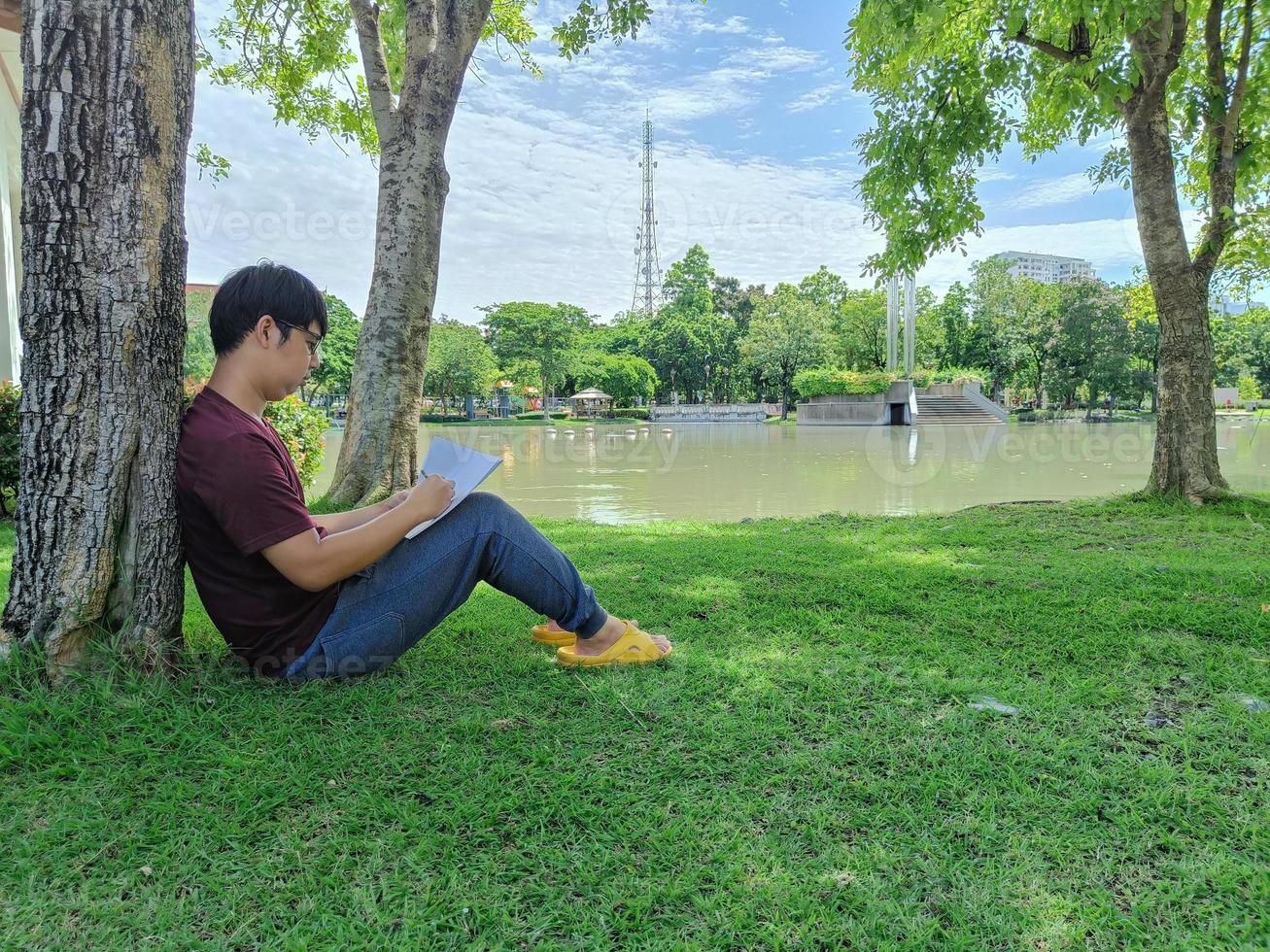 ung asiatisk man med svart hår sitter under ett träd och håller i en bok. stress och ångest. vad tänker du i sommarparkstaden foto