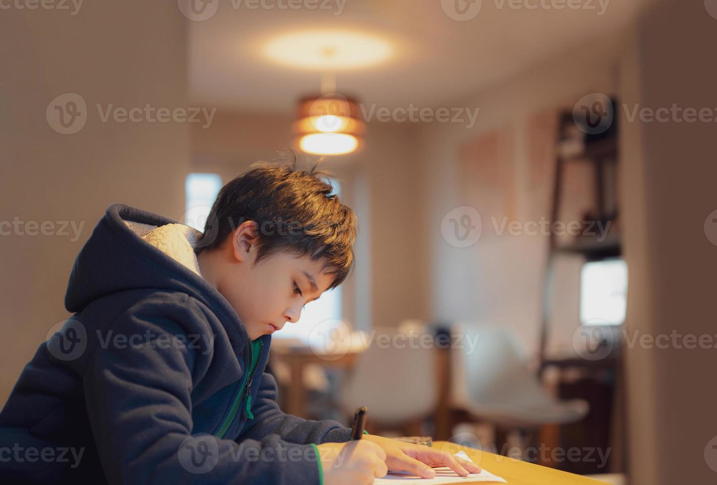 barn som sitter på bordet och gör läxor, barnpojke håller svart penna och skriver på vitt papper, ung pojke som tränar engelska ord hemma. grundskola och hemundervisning, distansundervisning koncept foto