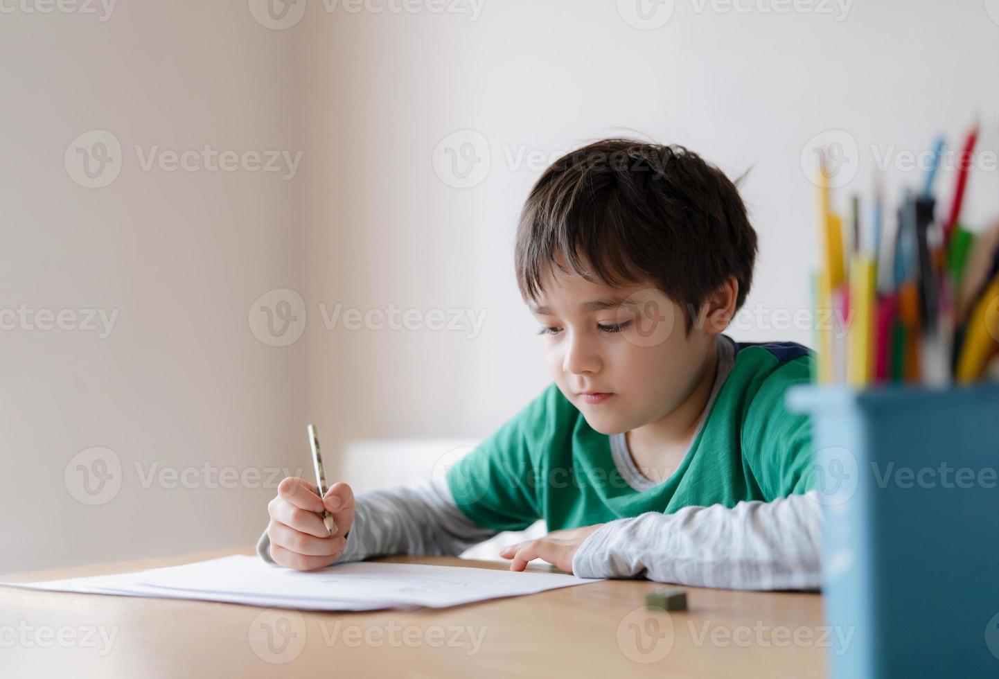 glad pojke som använder blyertsteckning eller skissar på papper, porträttbarn som sitter på bordet och gör läxor, barn tycker om konst och hantverksaktivitet hemma, utbildningskoncept foto