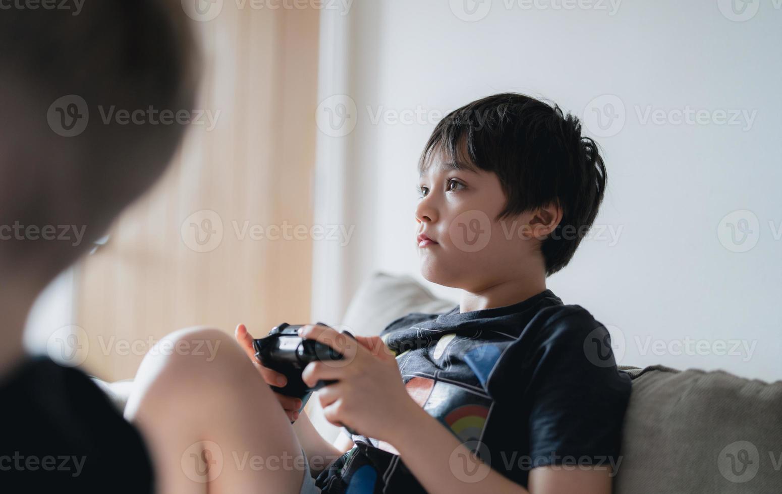 filmiskt porträtt ung pojke som spelar videospel online på internet, barn tittar upp och håller spelkontrollerande karaktärer med joystick, barn har roligt och umgås hemma foto