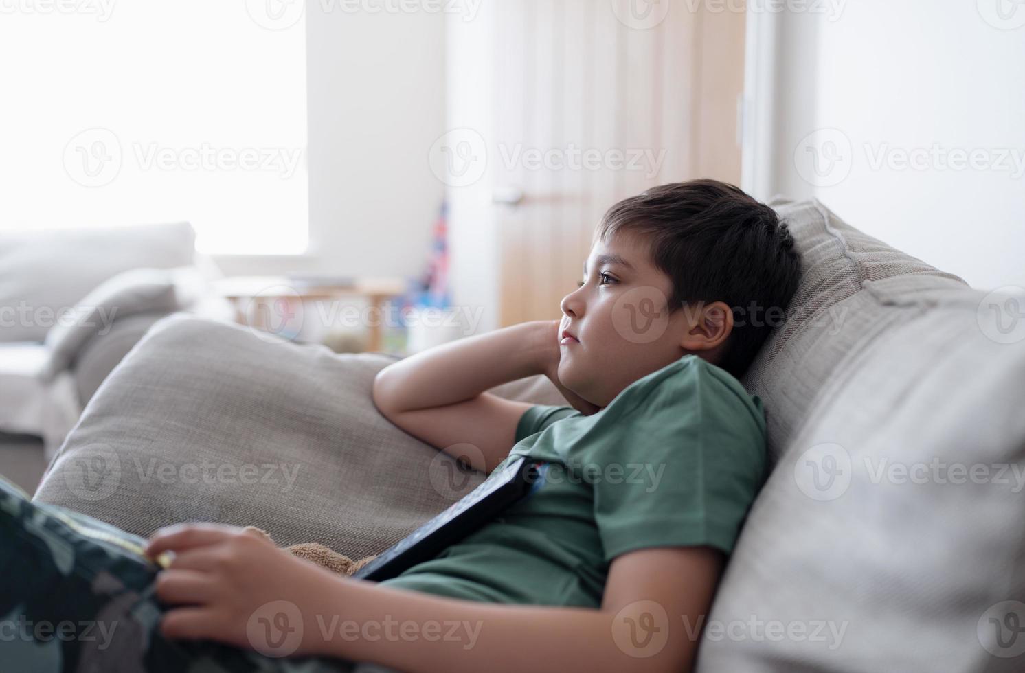 barn med fjärrkontroll och tittar upp med ett nyfiket ansikte, ung pojke som sitter i soffan och tittar på tecknad film på tv, porträtt av ett barn som ligger ner på soffan och kopplar av i vardagsrummet efter hemkomsten från skolan. foto