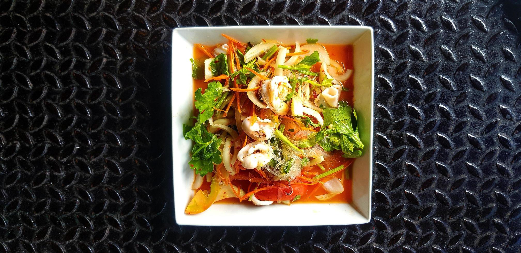 thai kryddig skaldjurssallad med glasnudlar, bläckfisk, räkor, skivad tomat, lök, morot och selleri på vit skål eller tallrik. asiatisk och berömd mat på svart bakgrund eller tapet av rostfritt stål. foto