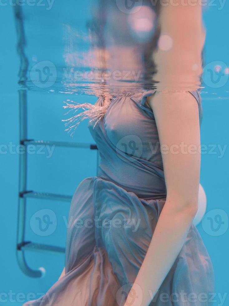 surrealistisk konstporträtt av ung kvinna i grå klänning och pärlhalsduk under vattnet i poolen foto