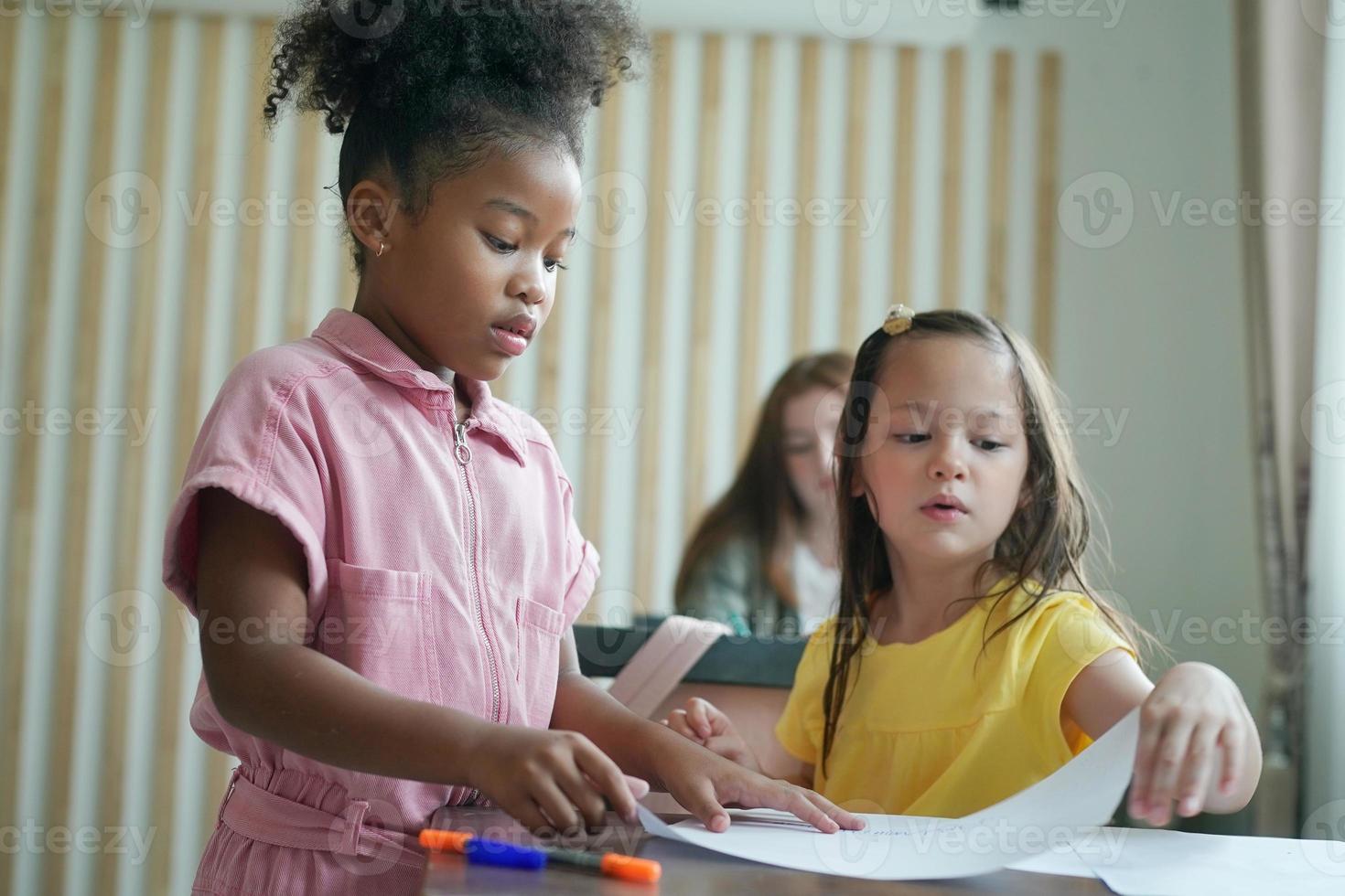 förskola flicka kid rita med färgpenna på vitt papper på bordet i klassrummet med vänner foto