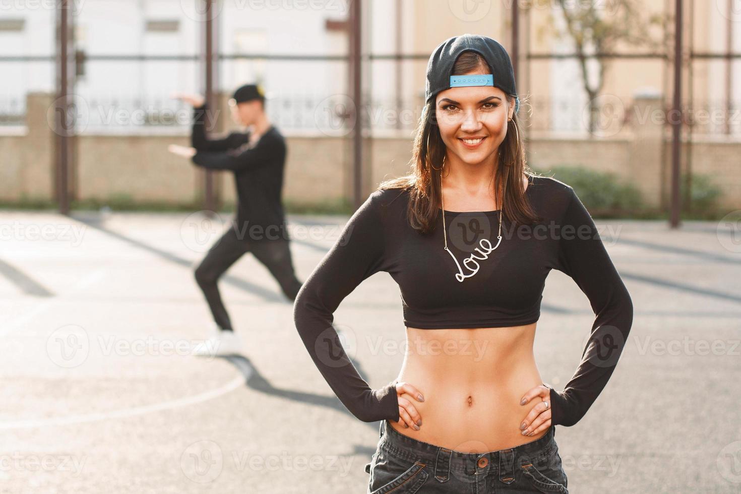 hip hop dansare kvinna i snygg svart klänning med kedja älskar och en keps. man står i en dansande pose i bakgrunden. foto