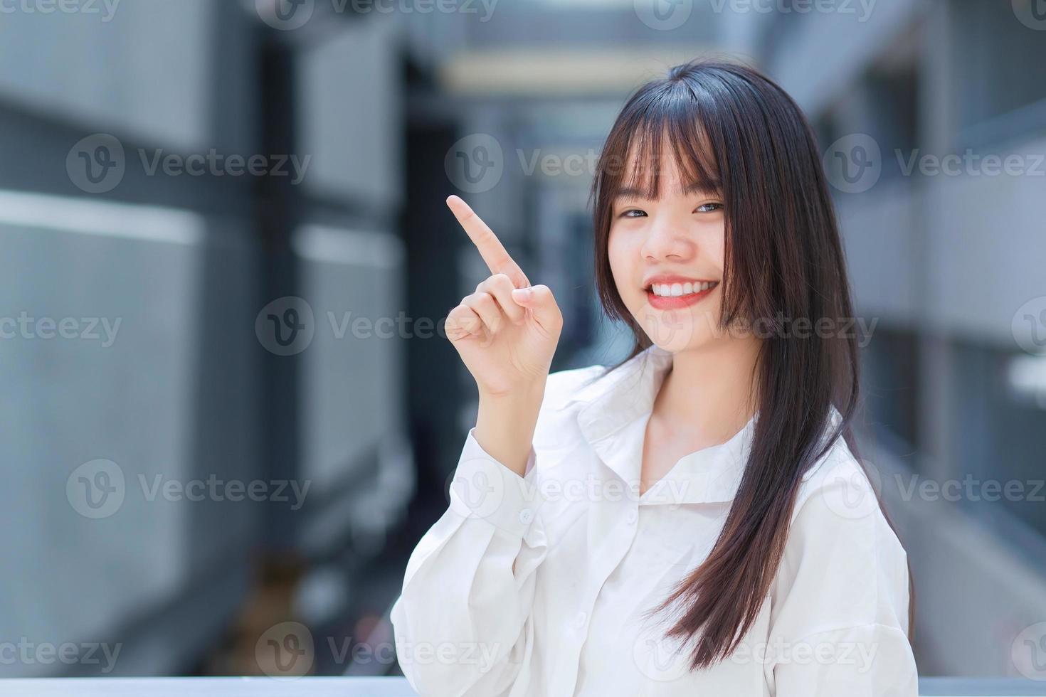 professionell ung asiatisk arbetande kvinna som bär vit skjorta pekar handen för att presentera något medans utomhus i staden med en kontorsbyggnad i bakgrunden. foto