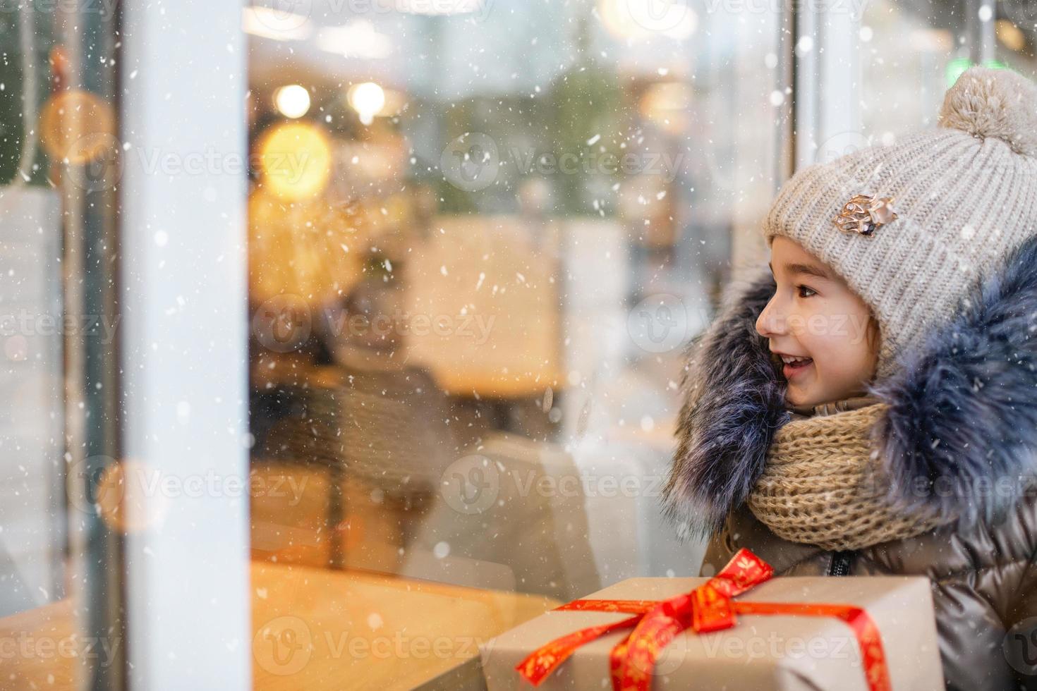 porträtt av glad tjej med en presentask för jul nära glasskyltfönstret på vintern med snö på festlig marknad med dekorationer och ljus. varma kläder, stickad mössa, halsduk och päls. kopieringsutrymme foto