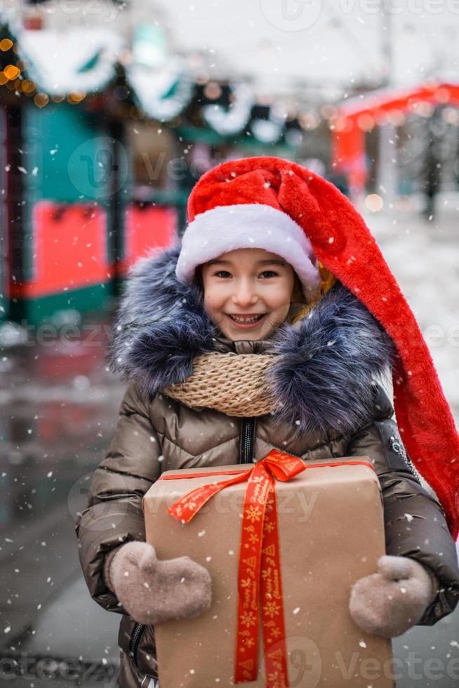 porträtt av glad flicka i tomtehatt med presentförpackning till jul på stadsgatan på vintern med snö på festlig marknad med dekorationer och fairy lights. varma kläder, stickad halsduk och päls. nyår foto