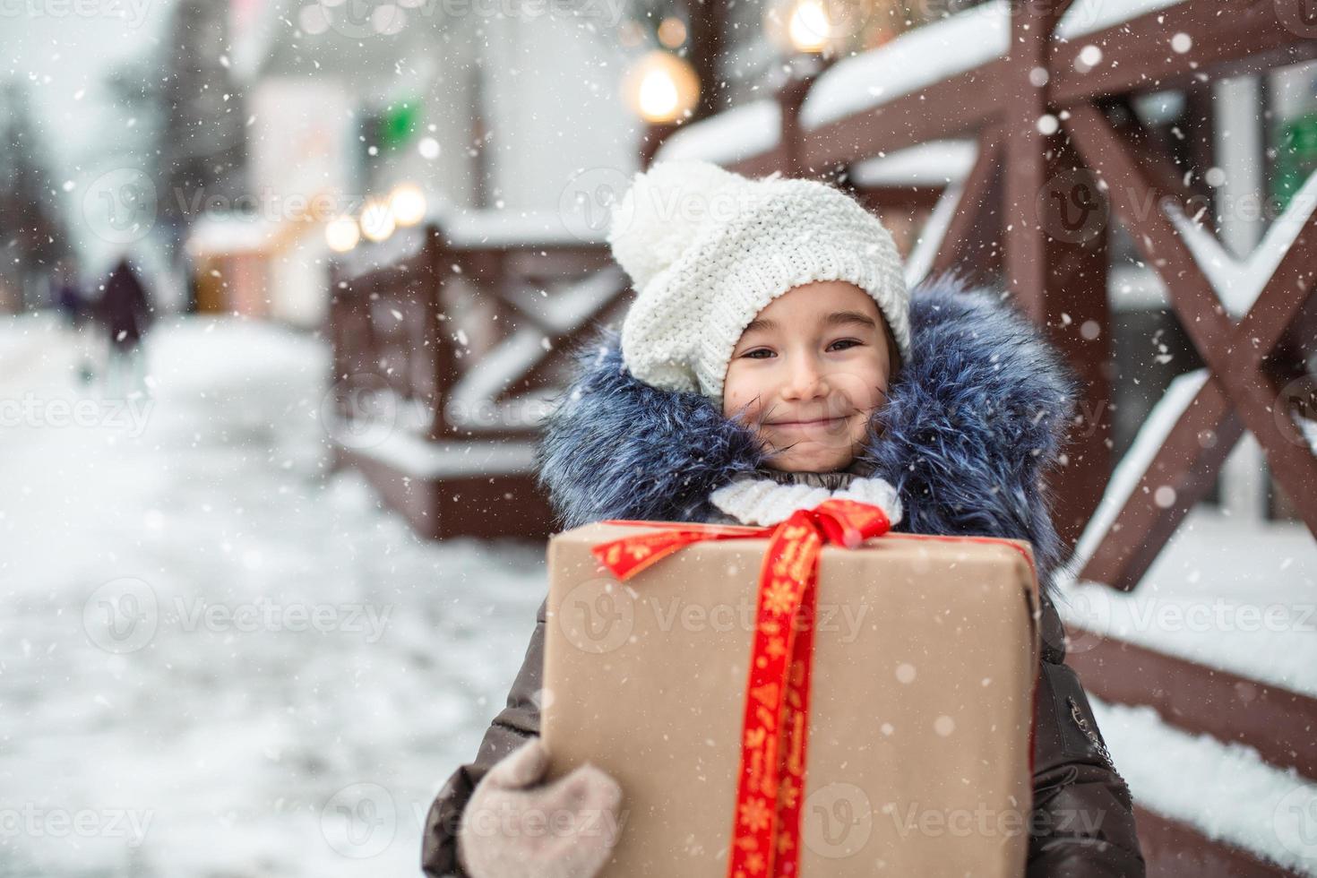 porträtt av glad tjej med en presentask till jul på en stadsgata på vintern med snö på en festlig marknad med dekorationer och älva ljus. varma kläder, stickad mössa, halsduk och päls. nyår foto