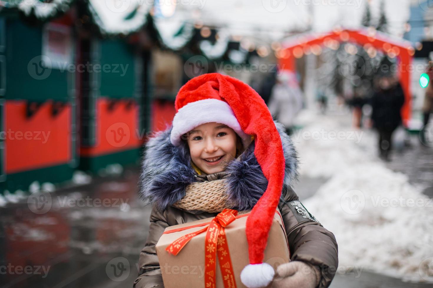 porträtt av glad flicka i tomtehatt med presentförpackning till jul på stadsgatan på vintern med snö på festlig marknad med dekorationer och fairy lights. varma kläder, stickad halsduk och päls. nyår foto