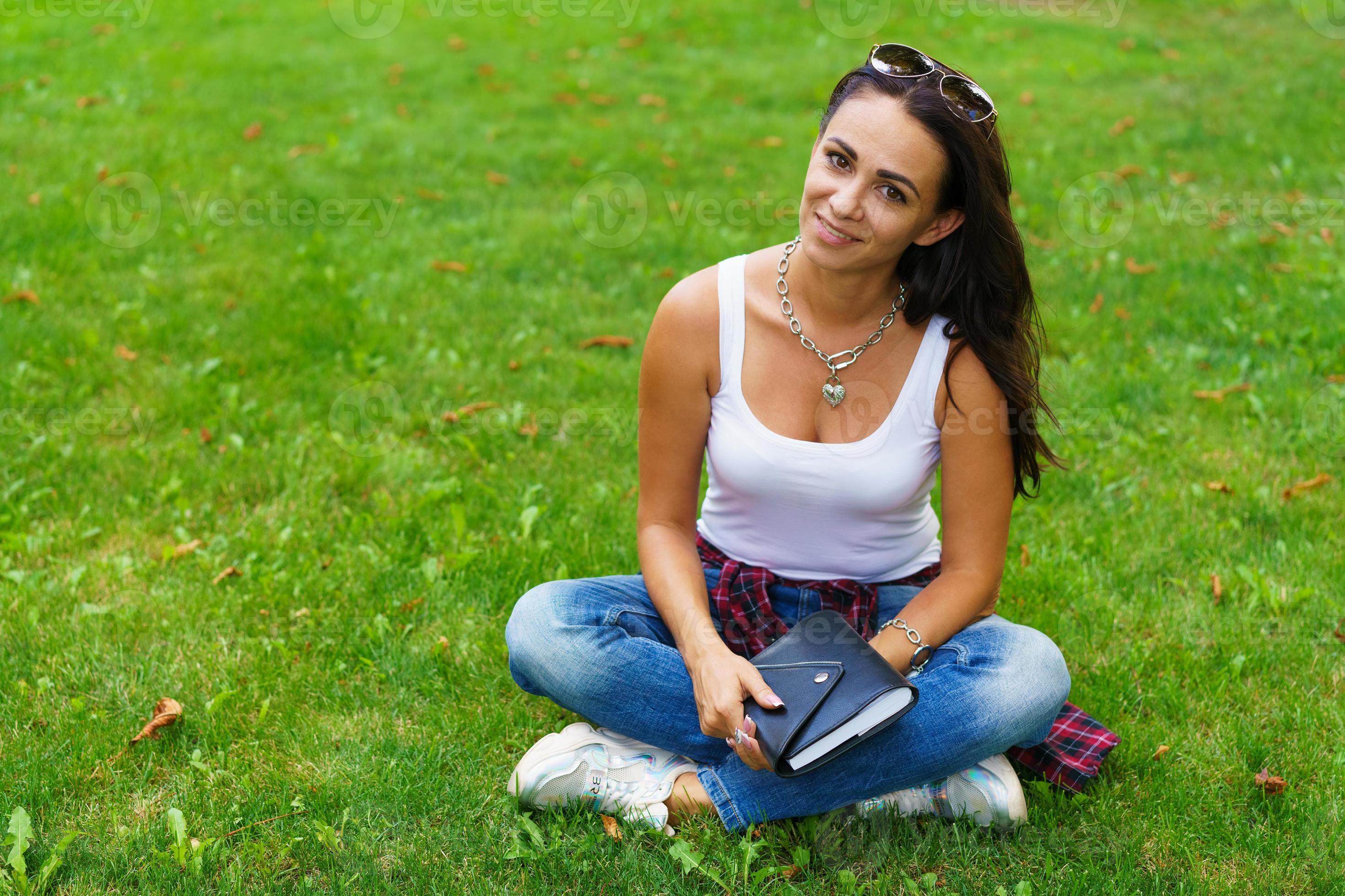 vacker flicka i vardagskläder sitter på gräset i universitetsparken med anteckningsblock foto