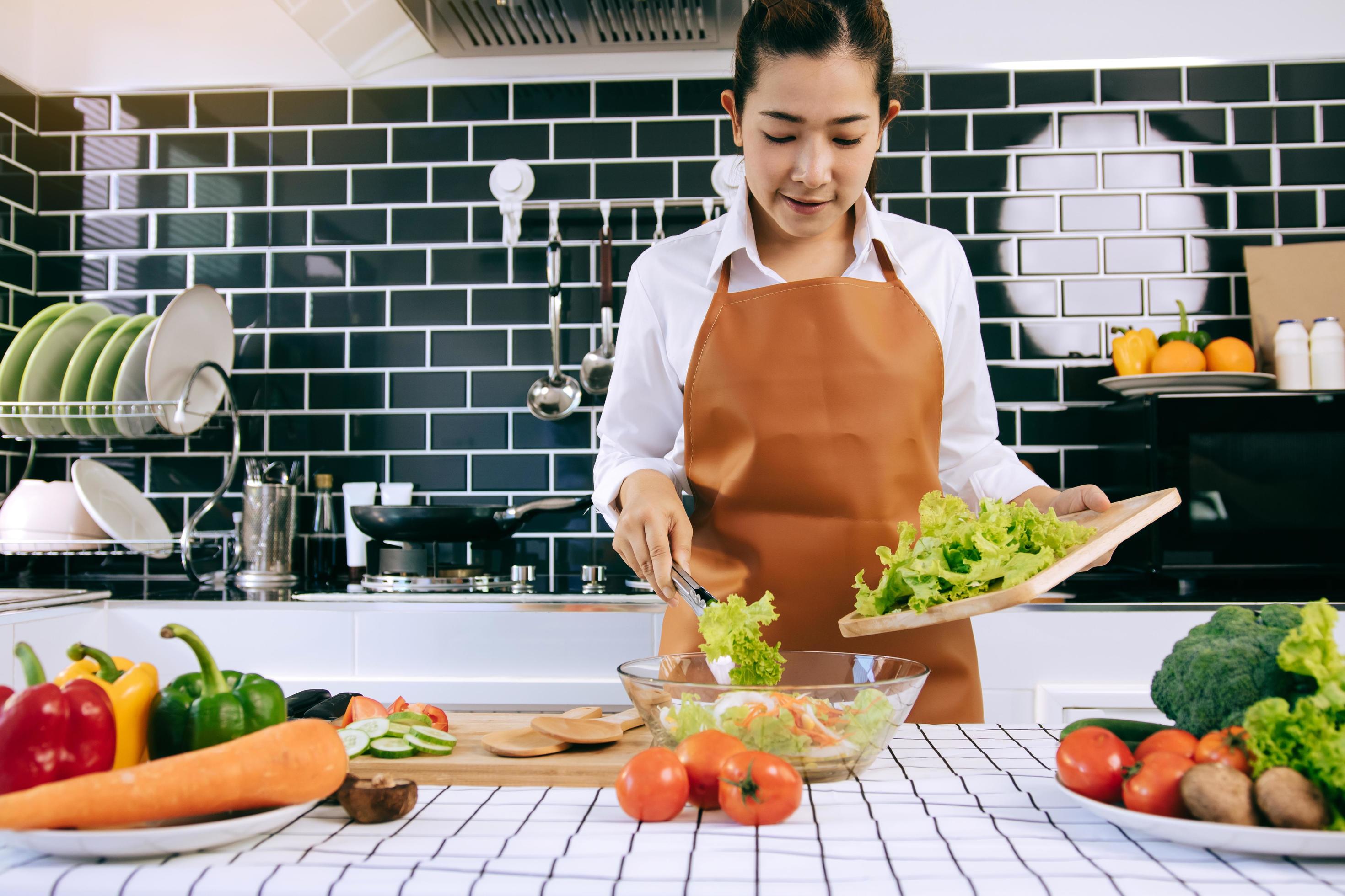 asiatisk hemmafru använder en tång för att ta salladen på träskärbrädan upp på salladskoppen i köksrummet. foto