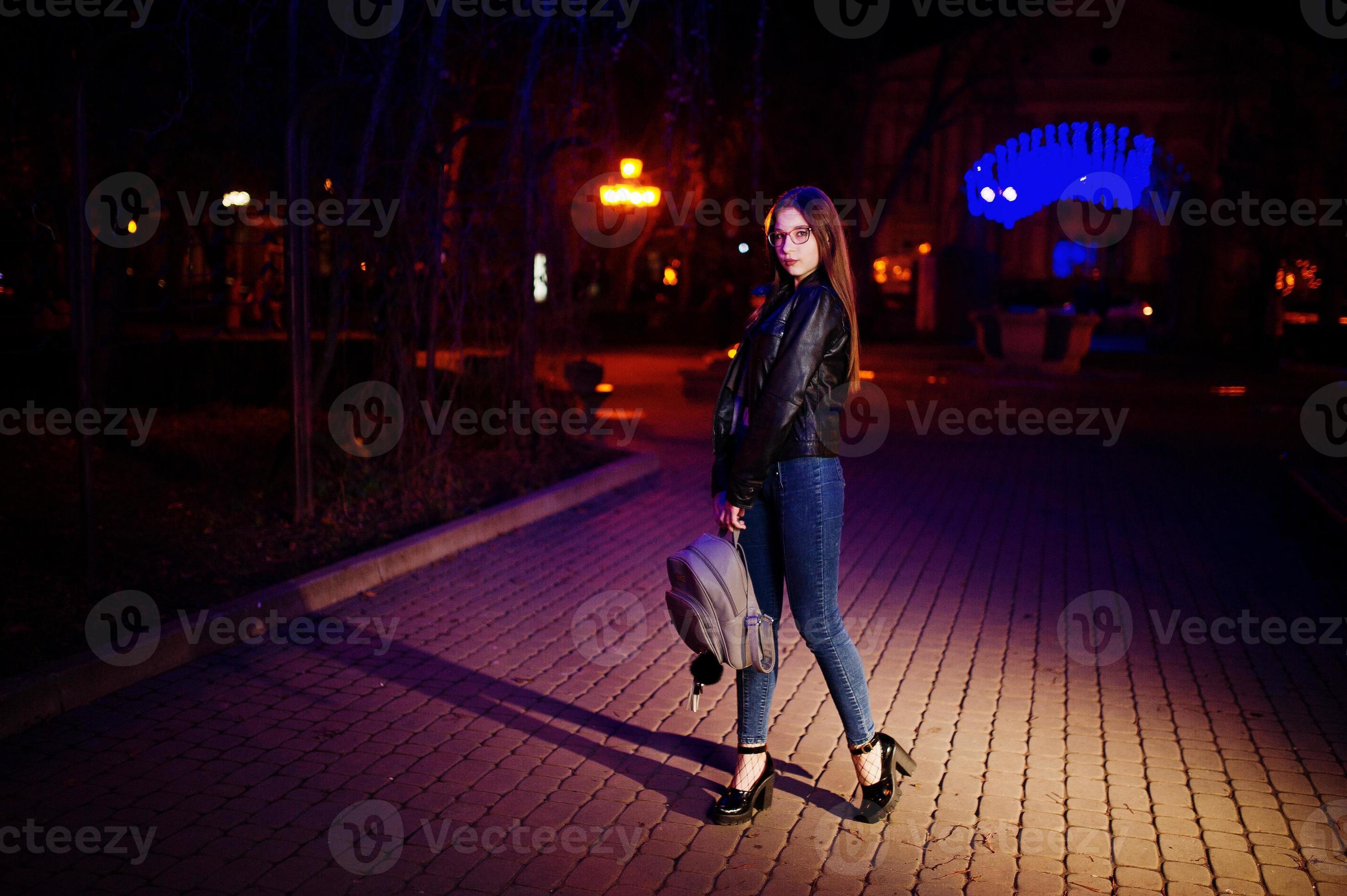 nattporträtt av flicka modell slitage på glasögon, jeans och skinnjacka,  med ryggsäck i händerna, mot blått ljus krans av stadsgatan. 8675253 Stock  Photo