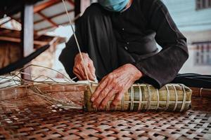 fokusera gammal kvinna med traditionell vietnamesisk klänning ao ba ba att göra, slå in tet tårta, det vietnamesiska månnyåret tet mat utomhus för händer. foto