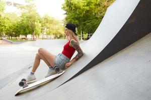 utomhusfoto av charmig smal ung blond kvinna som poserar över stuntparken en varm sommardag, lutad mot betongväggen medan hon har paus med ridbräda foto