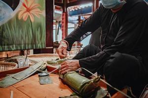 fokusera gammal kvinna med traditionell vietnamesisk klänning ao ba ba att göra, slå in tet tårta, det vietnamesiska månnyåret tet mat utomhus för händer. foto