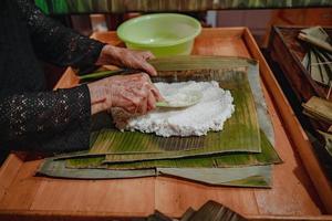 fokusera gammal kvinna med traditionell vietnamesisk klänning ao ba ba att göra, slå in tet tårta, det vietnamesiska månnyåret tet mat utomhus för händer. foto