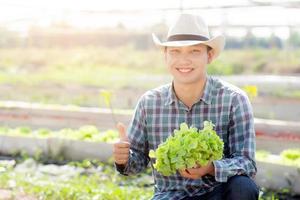 ung asiatisk man bonde håller och visar färsk organisk grön ek sallad och gest tummen upp i gård, producera och odling för skörd jordbruk grönsak med företag, hälsosam mat koncept. foto
